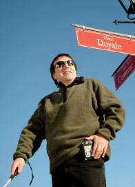 Photo of blind or low-vision male walking with white cane in one hand and Trekker in the other.